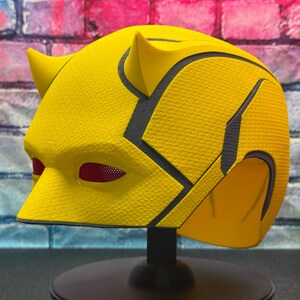 Puede incluir: Un casco de superh&eacute;roe amarillo con detalles negros y cubiertas oculares de malla roja. El casco tiene una superficie texturizada y se muestra en un soporte negro. El dise&ntilde;o incluye cuernos puntiagudos en la parte superior.