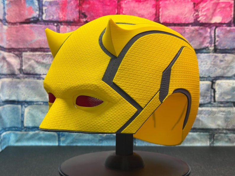 Puede incluir: Un casco de superh&eacute;roe amarillo con detalles negros y cubiertas oculares de malla roja. El casco tiene una superficie texturizada y se muestra en un soporte negro. El dise&ntilde;o incluye cuernos puntiagudos en la parte superior.