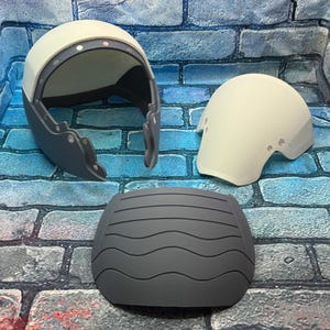 May include: Three-piece set of protective gear, including a helmet, elbow pad, and a chest protector. The helmet is white and gray, with a curved design. The elbow pad is white, and the chest protector is gray with a wavy pattern.