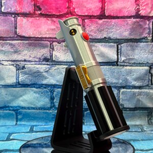 Pu&ograve; includere: Una replica di spada laser argento e nera con un pulsante rosso e dettagli dorati, esposta su un supporto nero. La spada laser &egrave; angolata contro un muro di mattoni con sfumature blu e rosa.