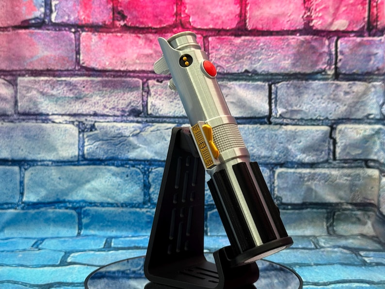 Pu&ograve; includere: Una replica di spada laser argento e nera con un pulsante rosso e dettagli dorati, esposta su un supporto nero. La spada laser &egrave; angolata contro un muro di mattoni con sfumature blu e rosa.