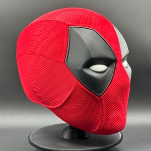 May include: A red and black Deadpool mask with a textured surface. The mask is on a black stand.