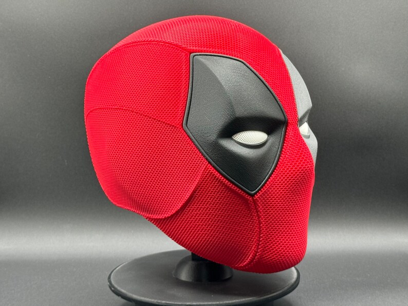 May include: A red and black Deadpool mask with a textured surface. The mask is on a black stand.