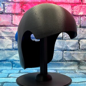 May include: A black helmet with a textured carbon fiber pattern and a smooth, rounded dome shape. The helmet features a large, circular blue accent on the side and is mounted on a black stand, displaying a sleek, modern design with cutouts.