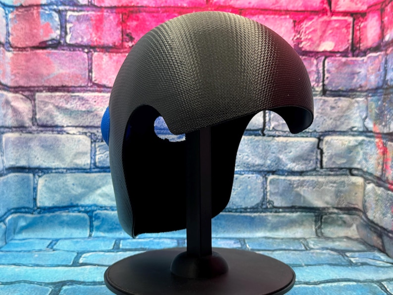 May include: A black helmet with a textured carbon fiber pattern and a smooth, rounded dome shape. The helmet features a large, circular blue accent on the side and is mounted on a black stand, displaying a sleek, modern design with cutouts.