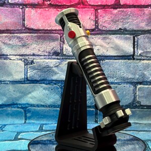 May include: A silver and black lightsaber prop with a red and yellow button detail, displayed on a black stand. The lightsaber has a ribbed black grip and silver accents. The background is a brick wall with blue and pink hues.