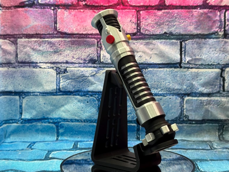 May include: A silver and black lightsaber prop with a red and yellow button detail, displayed on a black stand. The lightsaber has a ribbed black grip and silver accents. The background is a brick wall with blue and pink hues.