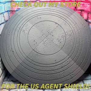May include: A gray, circular shield with concentric grooves and a textured surface. The shield has a central cross pattern and the text "CHECK OUT MY STORE" and "FOR THE US AGENT SHIELD!" in yellow.