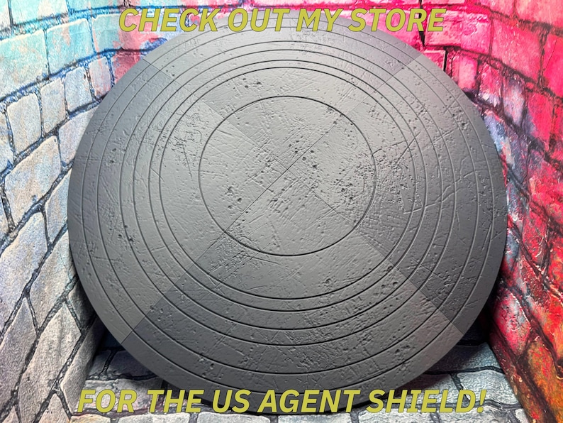 May include: A gray, circular shield with concentric grooves and a textured surface. The shield has a central cross pattern and the text "CHECK OUT MY STORE" and "FOR THE US AGENT SHIELD!" in yellow.