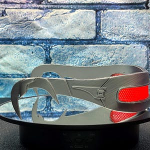 May include: A silver-colored, stylized headpiece with a curved design and sharp, claw-like extensions. The headpiece features two red, rectangular accents. The object is displayed on a mirrored surface against a blue brick background.