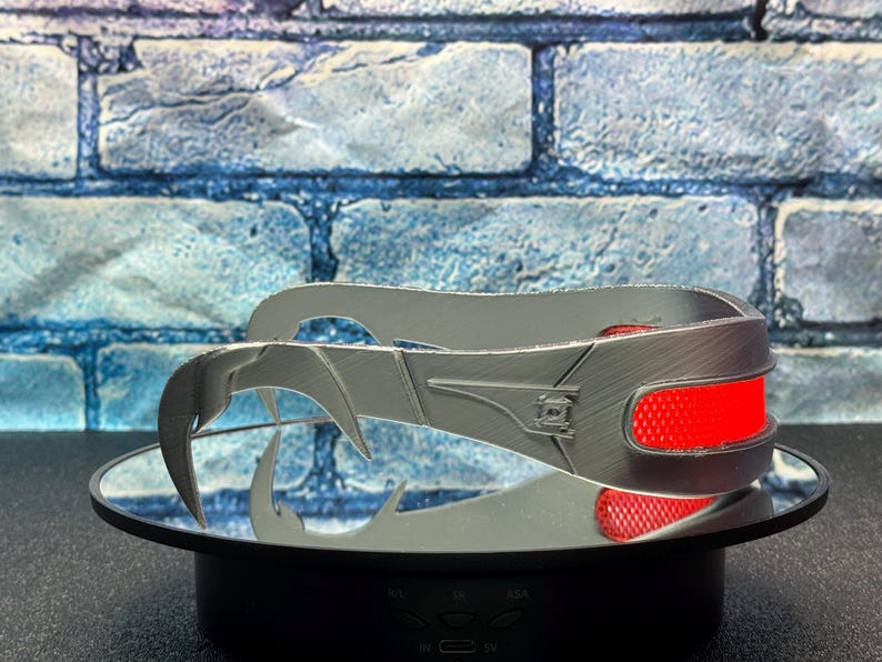 May include: A silver-colored, stylized headpiece with a curved design and sharp, claw-like extensions. The headpiece features two red, rectangular accents. The object is displayed on a mirrored surface against a blue brick background.
