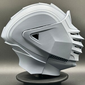 Puede incluir: Un casco gris impreso en 3D con una visera y un dise&ntilde;o detallado. El casco est&aacute; en un soporte negro.
