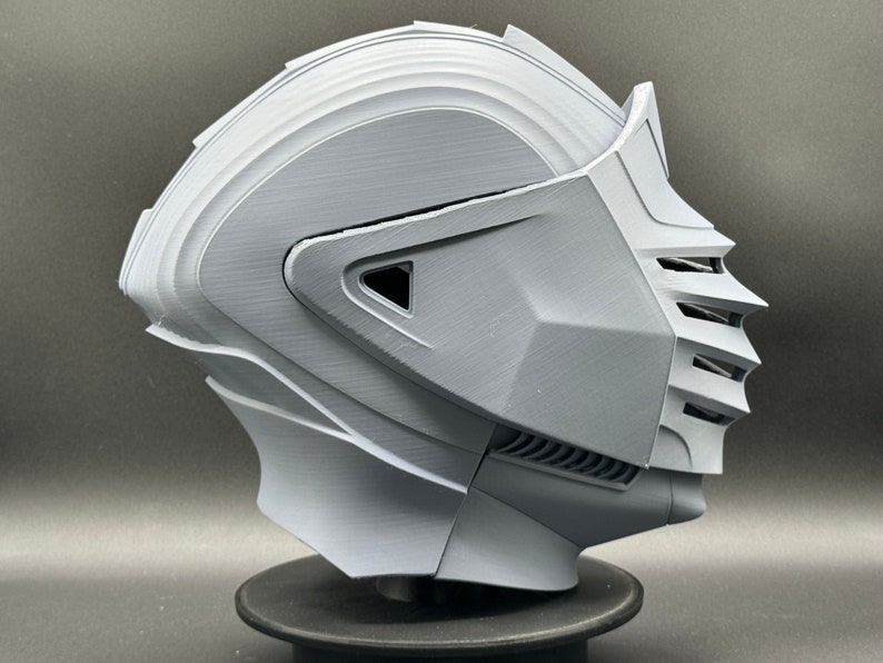 Puede incluir: Un casco gris impreso en 3D con una visera y un dise&ntilde;o detallado. El casco est&aacute; en un soporte negro.