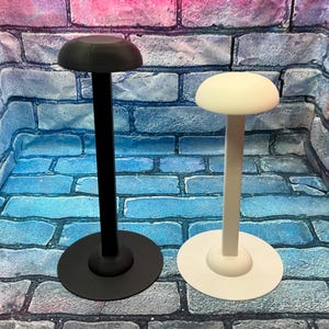 May include: Two hat stands, one black and one white, are displayed against a brick wall backdrop. Each stand features a circular base, a slender stem, and a rounded top, designed to hold hats. The black stand is on the left, and the white stand is on the right.