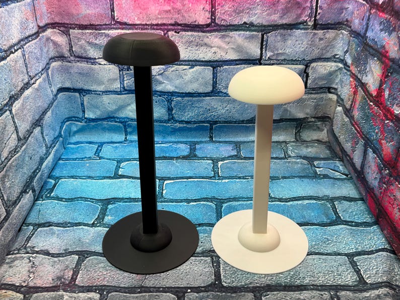May include: Two hat stands, one black and one white, are displayed against a brick wall backdrop. Each stand features a circular base, a slender stem, and a rounded top, designed to hold hats. The black stand is on the left, and the white stand is on the right.