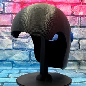 May include: A black helmet with a textured, carbon-fiber-like surface pattern and a smooth, curved dome shape. The helmet features a distinct cutout for the eye area and a small, circular blue accent piece attached to the side of the structure.