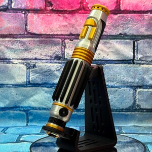 May include: A silver and gold lightsaber prop with black accents, resting on a black stand. The lightsaber features a cylindrical design with ribbed and panelled sections. The background is a brick wall with pink, blue, and purple hues.