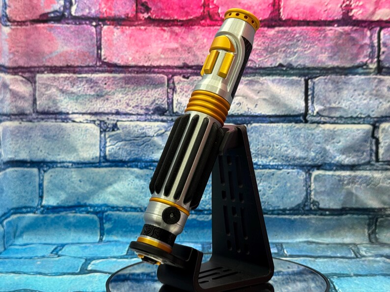May include: A silver and gold lightsaber prop with black accents, resting on a black stand. The lightsaber features a cylindrical design with ribbed and panelled sections. The background is a brick wall with pink, blue, and purple hues.