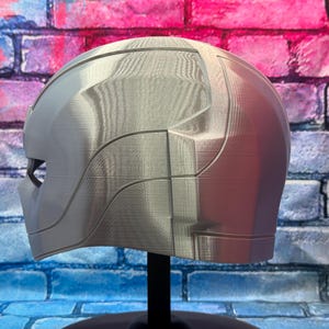 May include: A silver-colored helmet with a smooth, futuristic design. The helmet has a visor area and is displayed on a black stand. The surface has visible layer lines from the 3D printing process. The background is a brick wall.