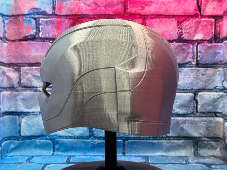 May include: A silver-colored helmet with a smooth, futuristic design. The helmet has a visor area and is displayed on a black stand. The surface has visible layer lines from the 3D printing process. The background is a brick wall.