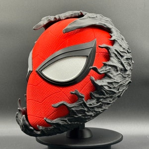 Puede incluir: Una m&aacute;scara de Spider-Man roja y negra con un dise&ntilde;o de simbionte Venom negro. La m&aacute;scara est&aacute; en un soporte negro.