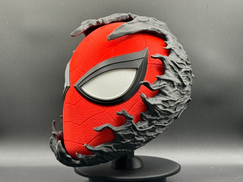 Puede incluir: Una m&aacute;scara de Spider-Man roja y negra con un dise&ntilde;o de simbionte Venom negro. La m&aacute;scara est&aacute; en un soporte negro.