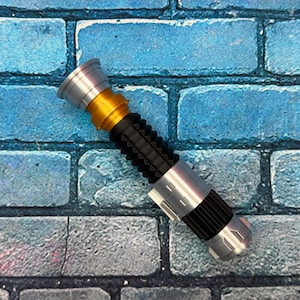 May include: A silver, gold, and black lightsaber prop. The hilt has a ribbed black grip, silver accents, and a gold midsection. The lightsaber is approximately 28 cm long.