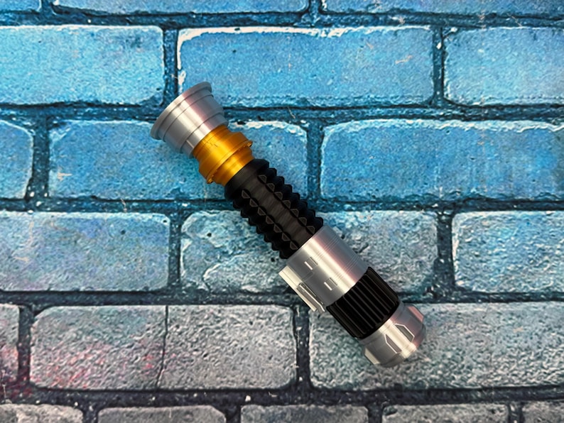 May include: A silver, gold, and black lightsaber prop. The hilt has a ribbed black grip, silver accents, and a gold midsection. The lightsaber is approximately 28 cm long.