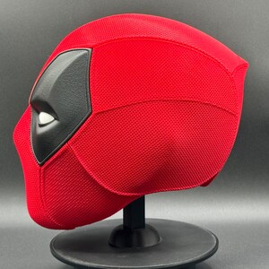 May include: A red and black Deadpool mask with a textured surface. The mask is on a black stand.