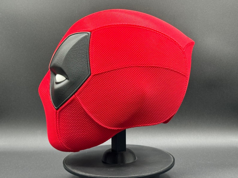 May include: A red and black Deadpool mask with a textured surface. The mask is on a black stand.