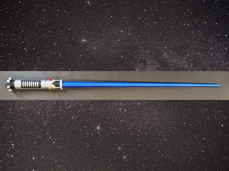 May include: A blue lightsaber prop with a silver and black handle. The prop is displayed against a starry background. The lightsaber has a long, cylindrical blue blade and detailed handle.
