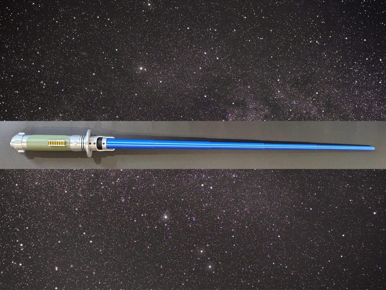Op de afbeelding: Een blauw lichtzwaard rekwisiet met een zilveren en groene handgreep. De handgreep heeft gedetailleerde zilveren accenten en een groen gedeelte met gele details. Het lichtzwaardblad is felblauw en de achtergrond is een sterrenhemel.