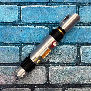 May include: A silver and black lightsaber prop with gold and red accents. The prop has a cylindrical design with detailed panels and a button. The lightsaber is set against a blue brick wall background.