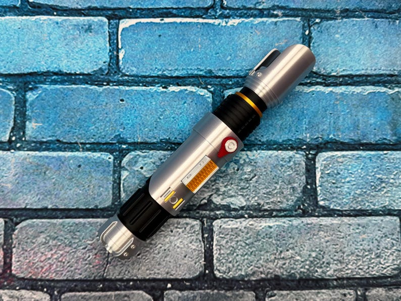 May include: A silver and black lightsaber prop with gold and red accents. The prop has a cylindrical design with detailed panels and a button. The lightsaber is set against a blue brick wall background.