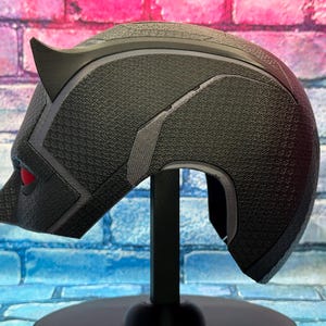 Puede incluir: Casco negro con superficie texturizada, con una oreja puntiaguda y detalle de ojo rojo. El casco se exhibe sobre un soporte negro contra un fondo de pared de ladrillo. El dise&ntilde;o del casco recuerda a un superh&eacute;roe.