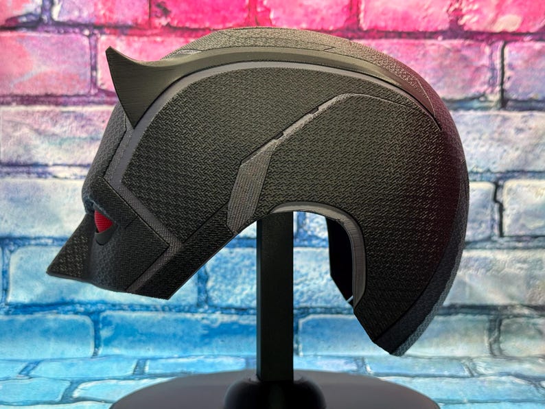 Puede incluir: Casco negro con superficie texturizada, con una oreja puntiaguda y detalle de ojo rojo. El casco se exhibe sobre un soporte negro contra un fondo de pared de ladrillo. El dise&ntilde;o del casco recuerda a un superh&eacute;roe.