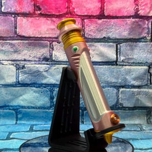 Op de afbeelding: Een roze en gouden lichtzwaard prop, rustend op een zwarte standaard. Het lichtzwaard heeft een metallic afwerking met zilveren accenten en een groene knop. De achtergrond is een bakstenen muur met roze en blauwe tinten.