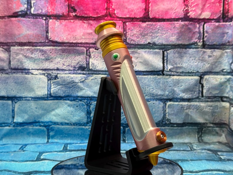 Op de afbeelding: Een roze en gouden lichtzwaard prop, rustend op een zwarte standaard. Het lichtzwaard heeft een metallic afwerking met zilveren accenten en een groene knop. De achtergrond is een bakstenen muur met roze en blauwe tinten.