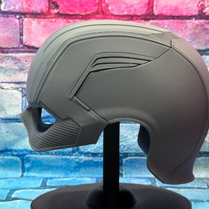 May include: A gray, futuristic-style helmet with a smooth, matte finish. The helmet features angular designs and a textured lower face guard. It is mounted on a black stand against a brick wall background.