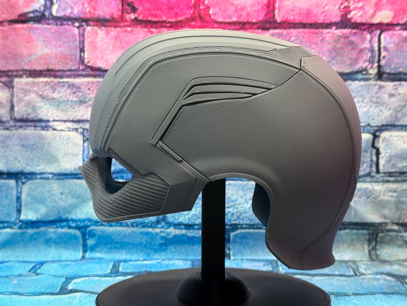 May include: A gray, futuristic-style helmet with a smooth, matte finish. The helmet features angular designs and a textured lower face guard. It is mounted on a black stand against a brick wall background.