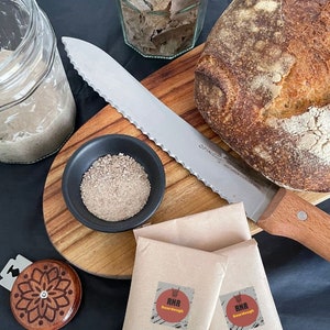 May include: A close-up of a sourdough bread loaf on a wooden chopping board with a knife, a small bowl of brown powder, and two brown paper bags with the text "RNR Sourdough" printed on them.