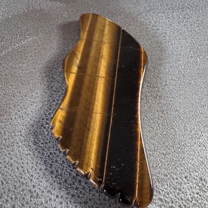 May include: A tiger's eye gemstone gua sha tool. The tool is shaped like a foot with a curved edge and a jagged edge. The stone has a striped pattern of brown and gold.