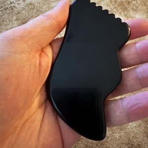 May include: A black gua sha tool, held in a hand, is shaped like a foot. The tool has a smooth, glossy surface and a scalloped edge. The background is a light brown, textured surface.