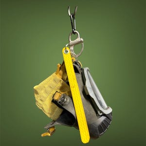 May include: A black and gray respirator mask with a clear visor, a yellow measuring stick, and a yellow mesh bag are suspended from a metal hook. The mask has a ribbed lower section and is attached to a silver carabiner.
