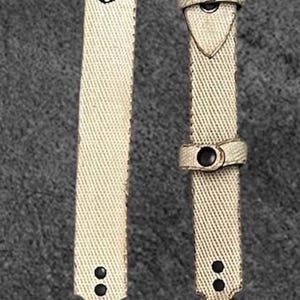 Könnte beinhalten: Cremefarbener Canvas-Gurt mit schwarzen Beschlägen, darunter eine Schnalle, Nieten und eine kleine Tasche. Das Wort "MOTES" ist vertikal auf dem Gurt aufgedruckt. Für eine Tasche oder Zubehör.