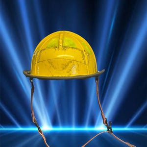 Könnte beinhalten: Ein gelber Feuerwehrhelm mit dunklem Rand und Kinnriemen. Der Helm hat ein leicht verwittertes Aussehen und verfügt über ein klares Visier. Der Hintergrund ist ein Blauton mit hellen Lichtstreifen.