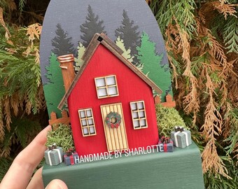 Può includere: Una decorazione da tavolo in legno raffigurante una casa rossa con tetto marrone, camino e una ghirlanda sulla porta. La scena include alberi verdi, muschio e piccoli pacchetti regalo. Il testo HANDMADE BY SHARLOTTE è stampato sulla base verde.