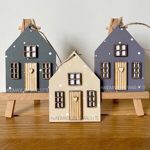 Pu&ograve; includere: Tre ornamenti in legno a forma di casa nei colori grigio scuro, beige chiaro e viola, decorati con pois bianchi, una porta centrale con un piccolo cuore e finestre. Ogni ornamento &egrave; dotato di un laccio in corda di juta.
