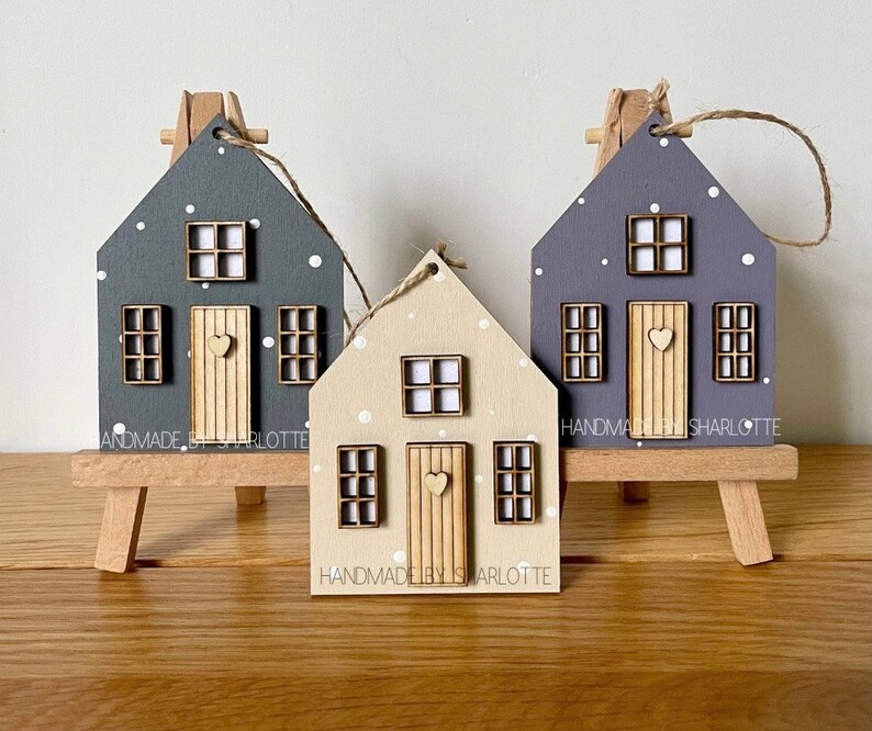 Pu&ograve; includere: Tre ornamenti in legno a forma di casa nei colori grigio scuro, beige chiaro e viola, decorati con pois bianchi, una porta centrale con un piccolo cuore e finestre. Ogni ornamento &egrave; dotato di un laccio in corda di juta.