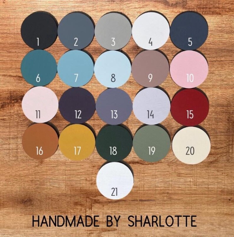 Pu&ograve; includere: Una collezione di ventuno campioni di vernice circolari in legno disposti in una griglia, ciascuno etichettato con un numero da 1 a 21 in testo bianco o nero. I colori includono nero, grigio, blu, rosa, rosso, giallo, verde e bianco. Testo: HANDMADE BY SHARLOTTE.